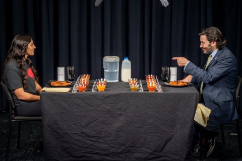 A man in a suit and tie gestures to a woman, both of whom are sitting in front of plates of chicken wings.
