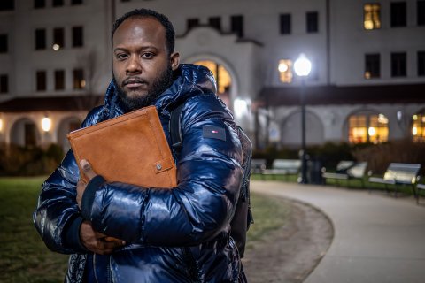 Horatio Wauchope , wearing a winter coat, stands outside on a campus walkway at night, holding a large folder to his chest in front of a lit academic building.