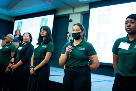 Five students in green polo shirts in front of projectors giving presentation