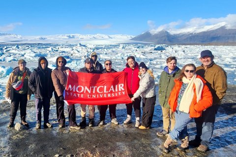 A group of Montclair students and professors stand together on a glacier in Iceland, holding a red Montclair State University banner. They are dressed in cold-weather gear, surrounded by a rugged, icy landscape. In the background, the glacier stretches into the distance, with patches of snow and exposed rock.