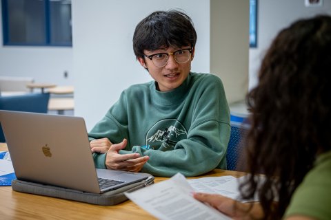 Student wearing green sweatshirt and glasses talks to another student across a table with his laptop open.