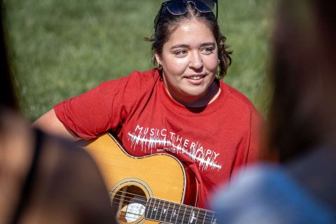 A student holds a guitar while seated in the grass.