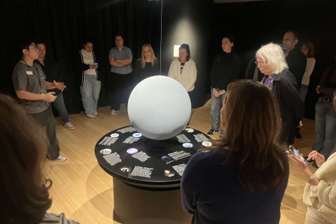 A group of people stand in a circle around an art exhibit and listen to a docent explain it