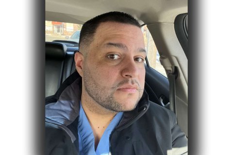 Headshot of a man wearing a blue shirt and a black jacket sitting inside a car