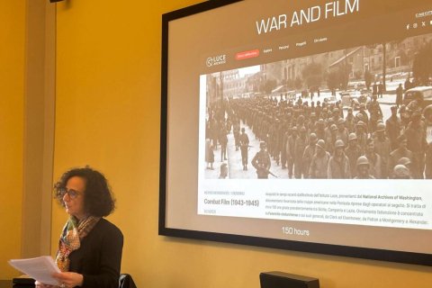 A college professor stands in front of a projector screen and delivers a presentation about "War and Film"