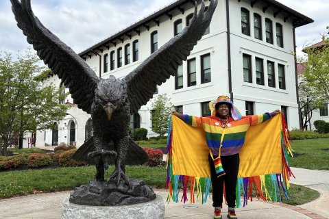 College student wearing a rainbow-colored fringed robe, rainbow sweater, colorful hat, and rainbow shoes, standing in front of a bronze statue of a Red Hawk.
