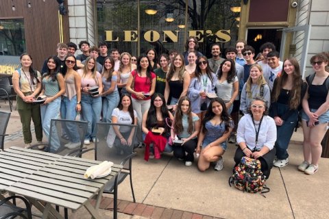 A large group of college students pose on a sidewalk outside an Italian restaurant