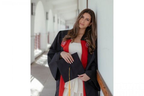A college student in a graduation robe holds a mortarboard and leans against the wall of a courtyard