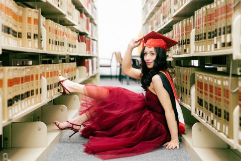 A college student dressed in a red cap and gown sits on the floor between shelves in a library