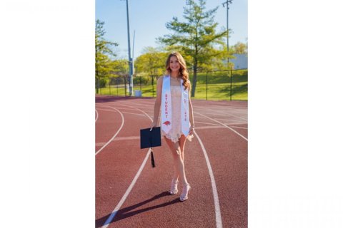 A college student stands holding a mortarboard on an outdoor running track