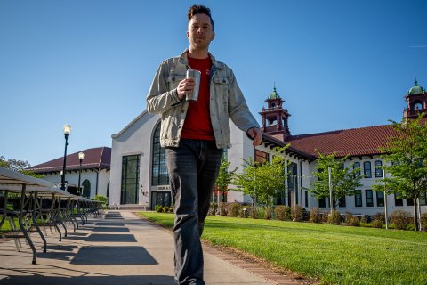 Casey Coleman walks along a campus pathway, holding a travel coffee mug, with long rows of empty folding tables on one side.