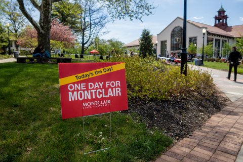 A red sign with while lettering says: One Day for Montclair. The campus is in background.