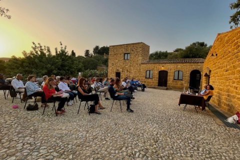 A woman sits at an outdoor table in a stone courtyard and speaks to an audience