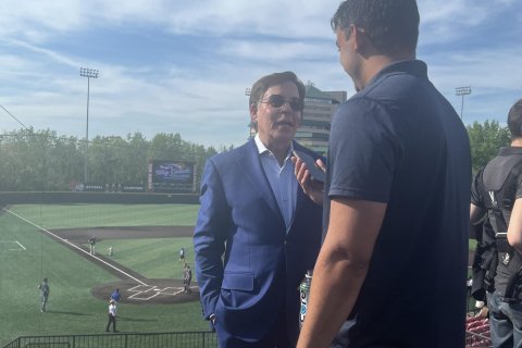 wo people standing outside in an empty sports stadium. The on in the foreground is interviewing the other person using is mobile phone.