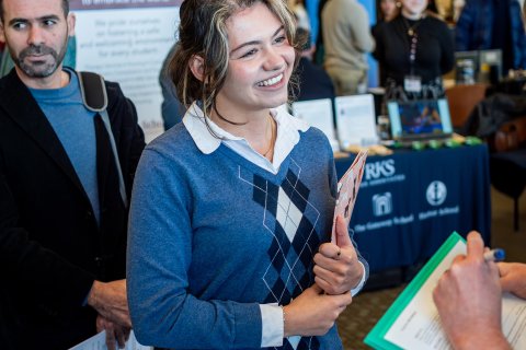 a woman smiles while talking to prospective employers at a career fair, holding papers in her hand