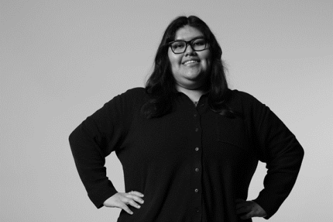 A black and white photo of a woman with long hair and glasses. She is smiling, standing and with her hands on her hips.