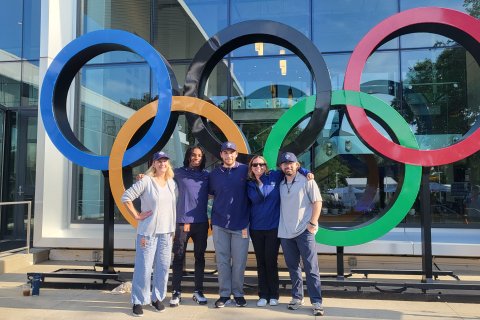 Five people standing and smiling in front of the Olympic Rings, which are large, colored circles.