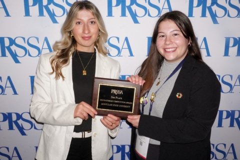 Two college students dressed professionally standing in front of a white backdrop with blue lettering. They are holding an award.