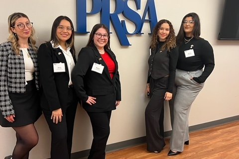 A group of five students standing and smiling with the letters P R S S A on the wall behind them.