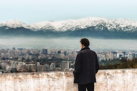 A man stands at a wall and looks over a cityscape with a large mountain range in the background