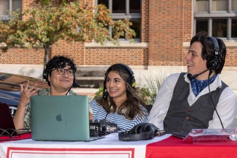 Three young students sitting outside at a table with a laptop in front of them. All of them are wearing headphones and are part of a live radio broadcast. They are smiling and happy.