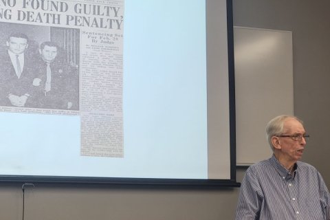 An old man stands in front of a projector screen and delivers a lecture