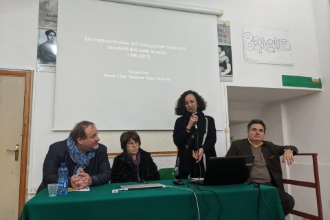 Four people stand at a presentation table and one stands and holds a microphone