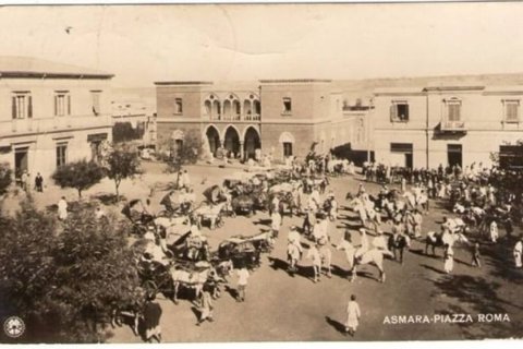 A sepia-toned image shows a busy town square in Asmara in Piazza Roma with horse-drawn carts, pedestrians, and surrounding buildings