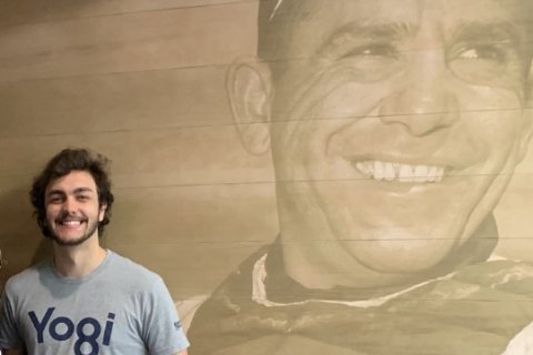 A college student stands in front of a wall decal of Yogi Berra from his playing days