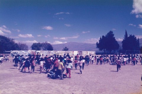 A large grouup of children of various ages run around an outdoor dirt field