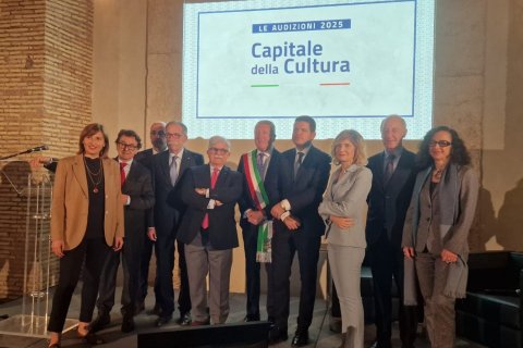 A group of people pose in front of a projector screen bearing the words "Le Audizioni 2025 Capitale della Cuultura""