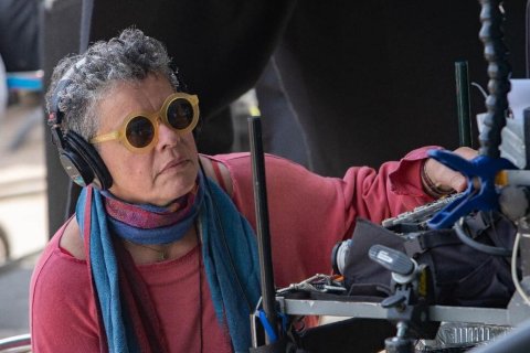A woman wears sunglasses and headphones and looks toward a screen