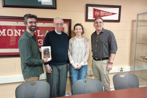 Four people stand inside a building with painted brick walls, one person holds up a book