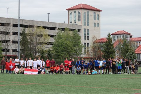 A large group of college students in soccer attire stand along the edge of a pitch and hold flags