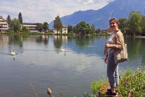 A woman stands at the edge of a lake with mountains and buildings across from her