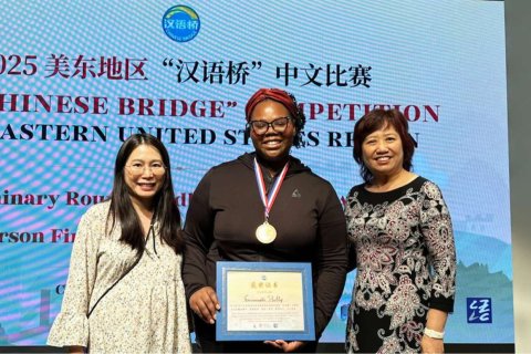 Three women pose for a photo in front of a projection screen, the woman in the middle wears a medal and holds a framed certificate