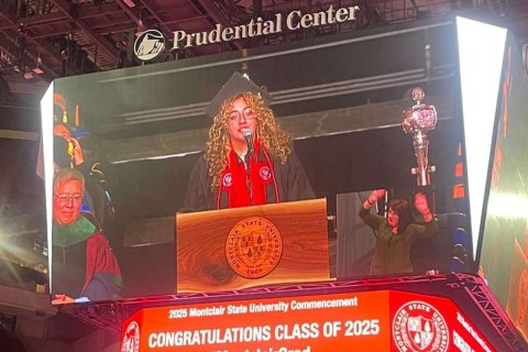 On a video board, a woman wearing graduation regalia speaks at a podium