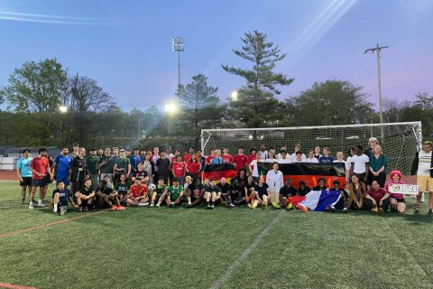 A group of college students and professors hold flags and pose in front of a goal on a soccer field