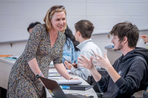 A woman leans on a desk and speaks to a college student 