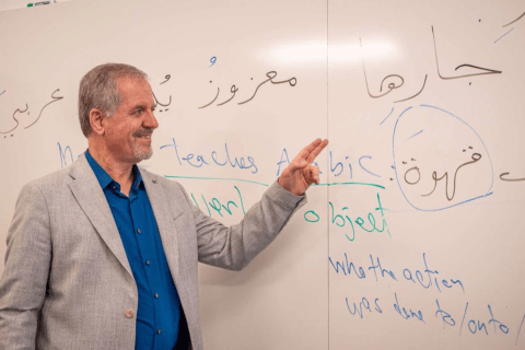 A man in a suit stands in front of a whiteboard and points to a piece of writing