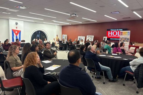People sit around several round tables in a conference room with a video board reading "Visiones de Cuba" and watch a presenter 