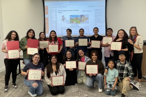 A group of people pose in two lines in front of a projector screen, nearly everyone holds up a framed paper certificate