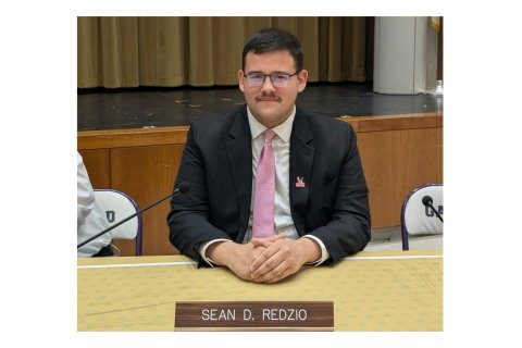 A man wears a suit and tie and sits at a desk with a name plate in front of it