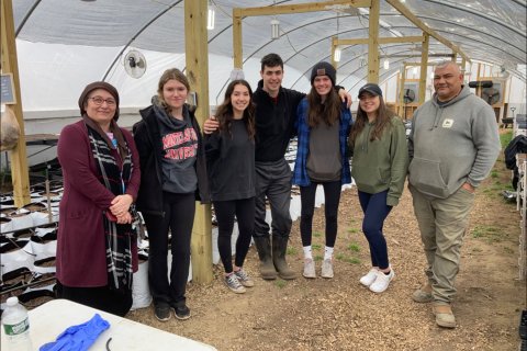 A group of people wearing cold weather attire stand in side a greenhouse and pose