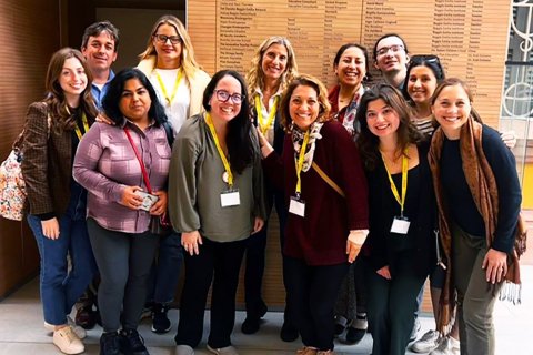 A group of people wear lanyards and pose in front of a wooden sign inside a building