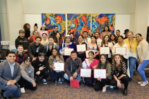 A large group of college students, some holding paper certificates, pose in front of an art installation