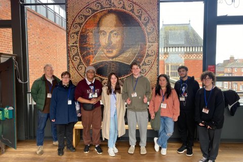 A group of people pose indoors in front of a tapestry of William Shakespeare's face.