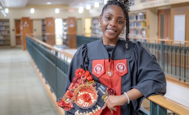 Lataiva Balmer, a psychology major and member of the first graduating class of Bloomfield College of Montclair State University, is shown in the college library wearing her cap and gown on the Bloomfield campus. She holds a decorated graduation cap.
