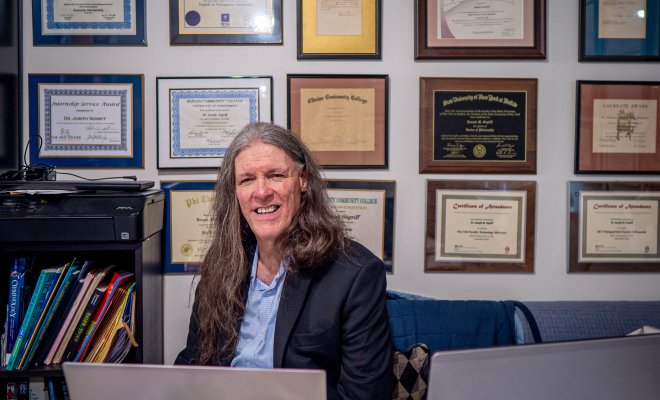 A man sits at his desk smiling with many diplomas on the wall behind him.