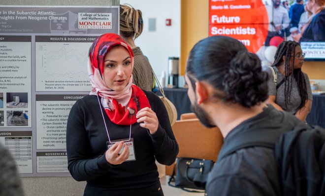 Busra Anil, a Montclair State University student wearing a red and white patterned hijab and black sweater, speaks with a guest during the Spring 2024 Mario M. Casabona Future Scientists Competition. She stands in front of her research poster on carbon sequestration, explaining her findings.